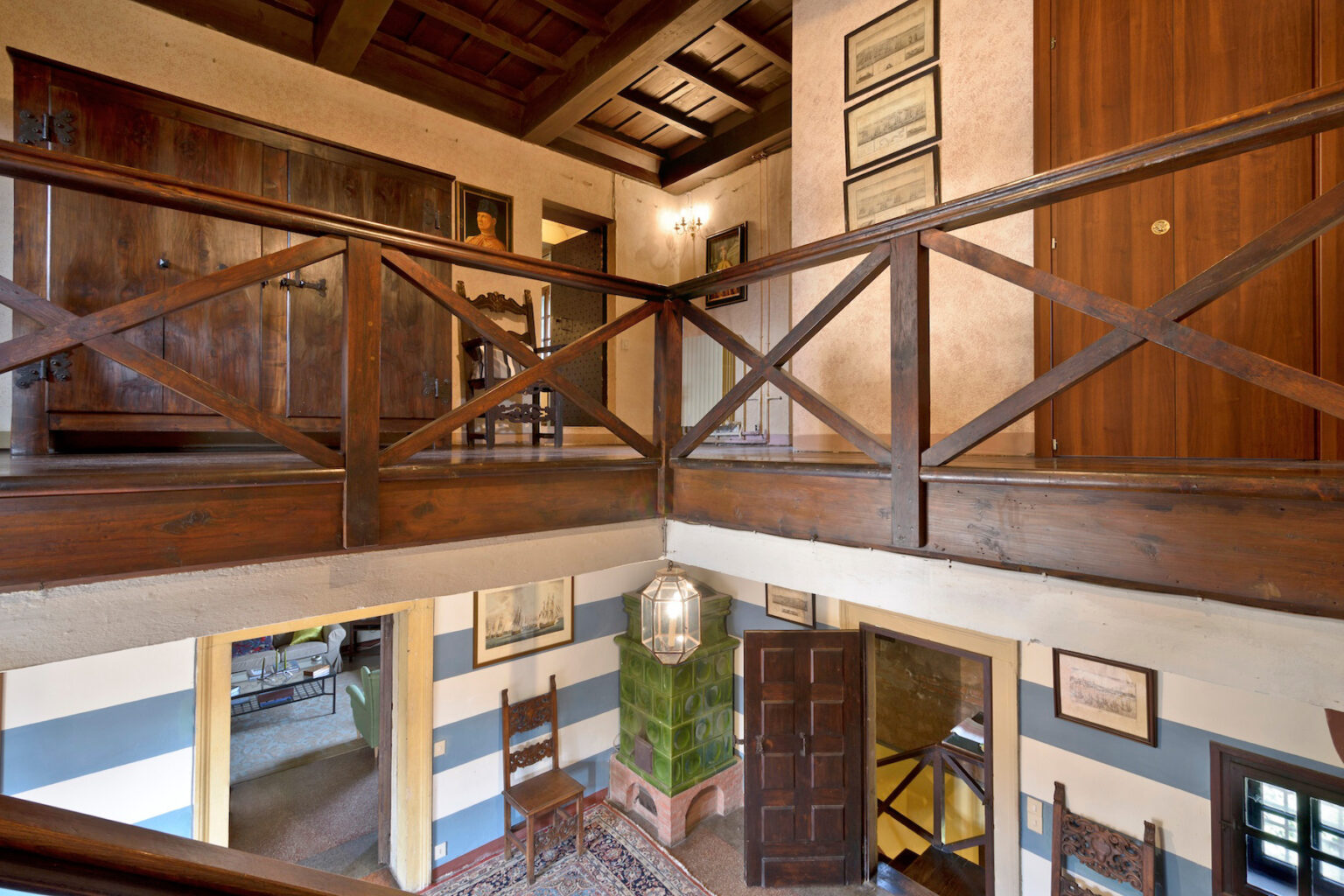 Interior view of a spacious, rustic home showcasing a wooden balcony with a crisscross railing, wooden doors, and framed artwork on the walls, illuminated by natural light. | Only on SopranoVillas