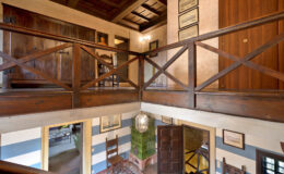 Interior view of a spacious, rustic home showcasing a wooden balcony with a crisscross railing, wooden doors, and framed artwork on the walls, illuminated by natural light.