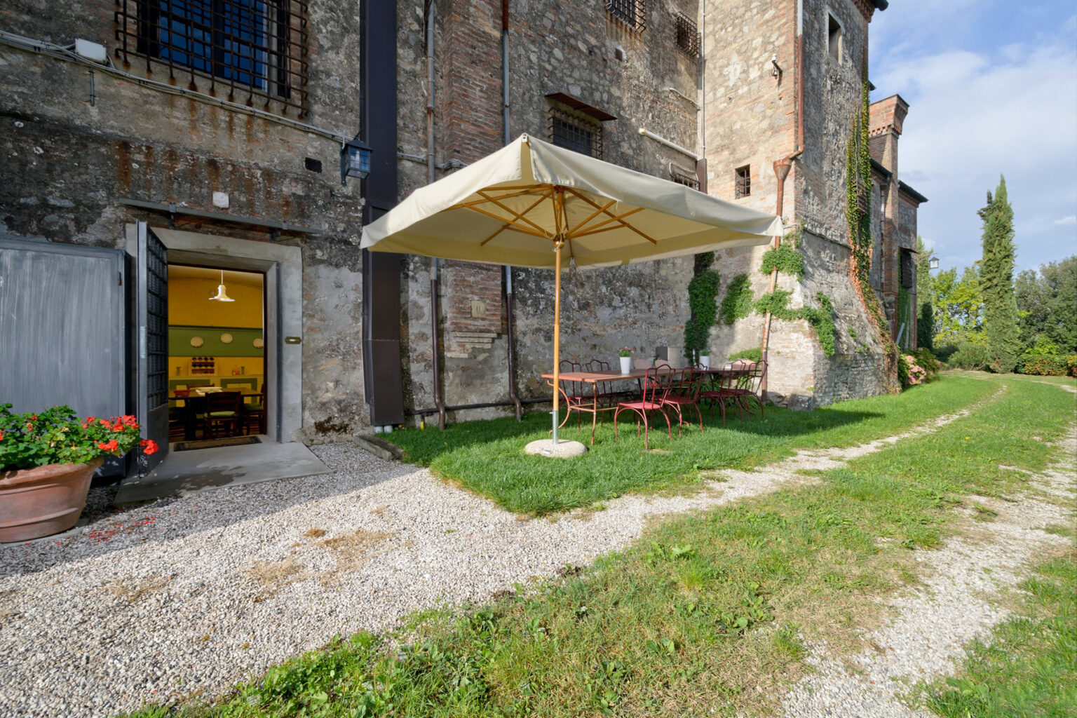 Outdoor seating area with a large umbrella next to a rustic building, featuring a gravel pathway and green lawn, ideal for relaxation and dining. | Only on SopranoVillas