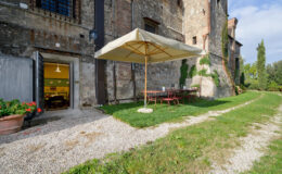 Outdoor seating area with a large umbrella next to a rustic building, featuring a gravel pathway and green lawn, ideal for relaxation and dining.