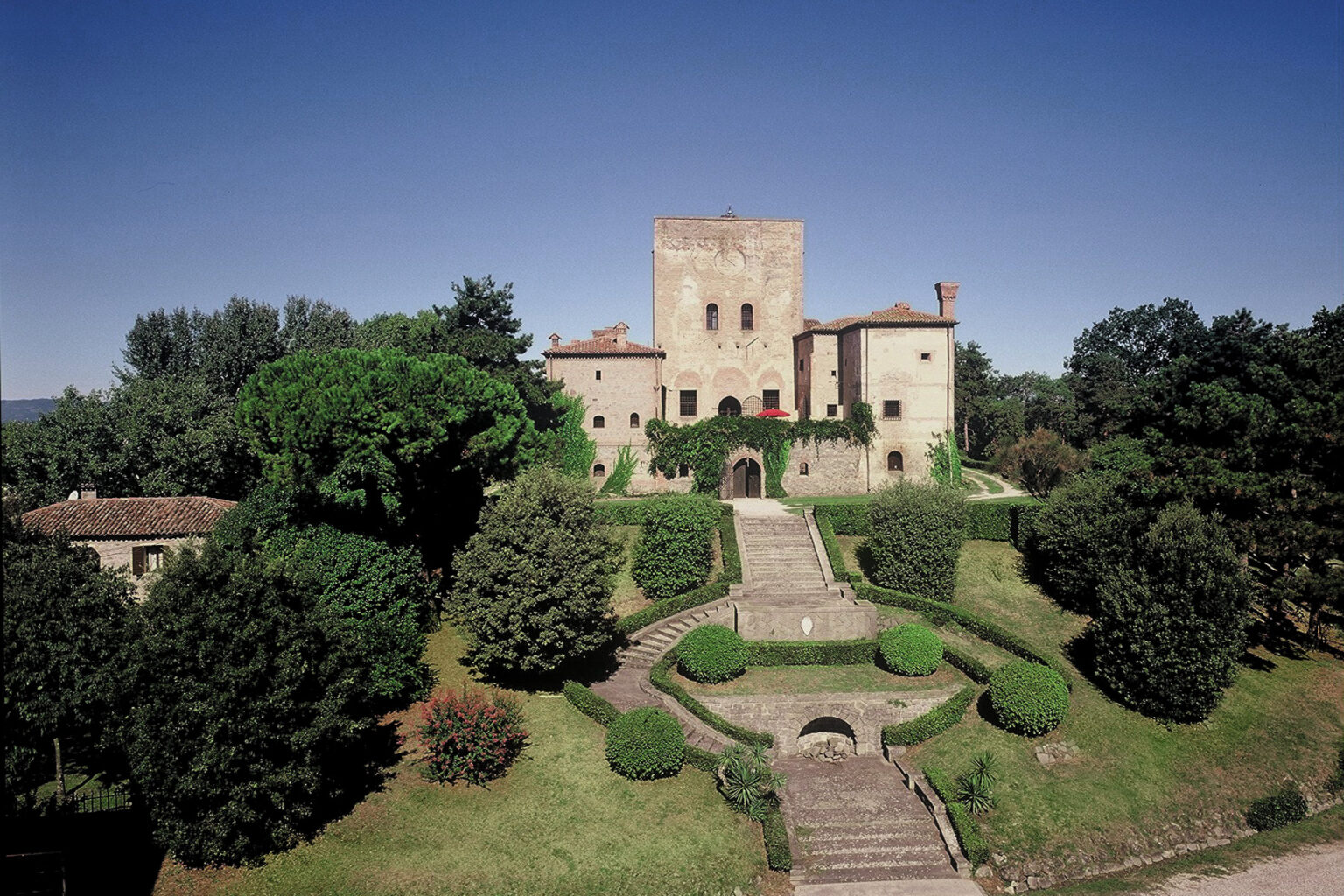 Historic castle surrounded by lush greenery and manicured gardens under a clear blue sky. The architectural features include a tall tower and stone walls, showcasing medieval design. Ideal for travel enthusiasts and history lovers. | Only on SopranoVillas
