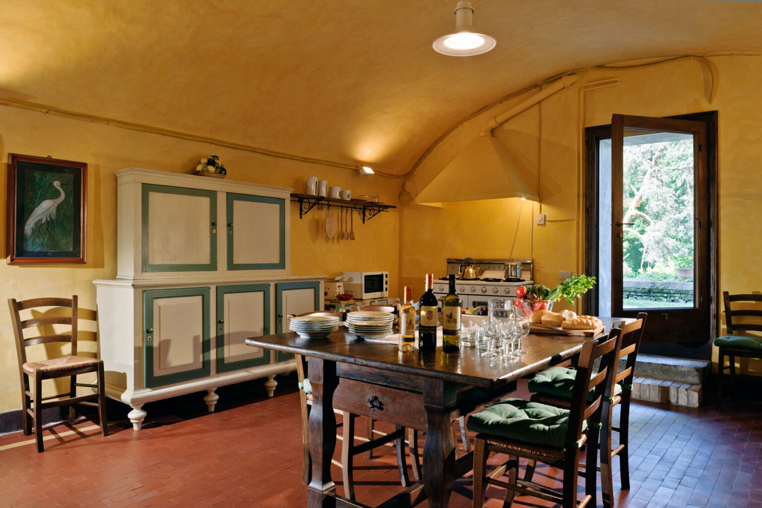 Cozy kitchen interior featuring a wooden dining table surrounded by chairs, a green and white cabinet, and a large window opening to a garden view. The warm yellow walls enhance the inviting atmosphere, perfect for family gatherings or casual dining. | Only on SopranoVillas