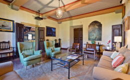 Cozy living room with green armchairs, a beige sofa, and a coffee table, featuring warm lighting and artwork on the walls. The room has a classic design with wooden accents and a welcoming atmosphere.