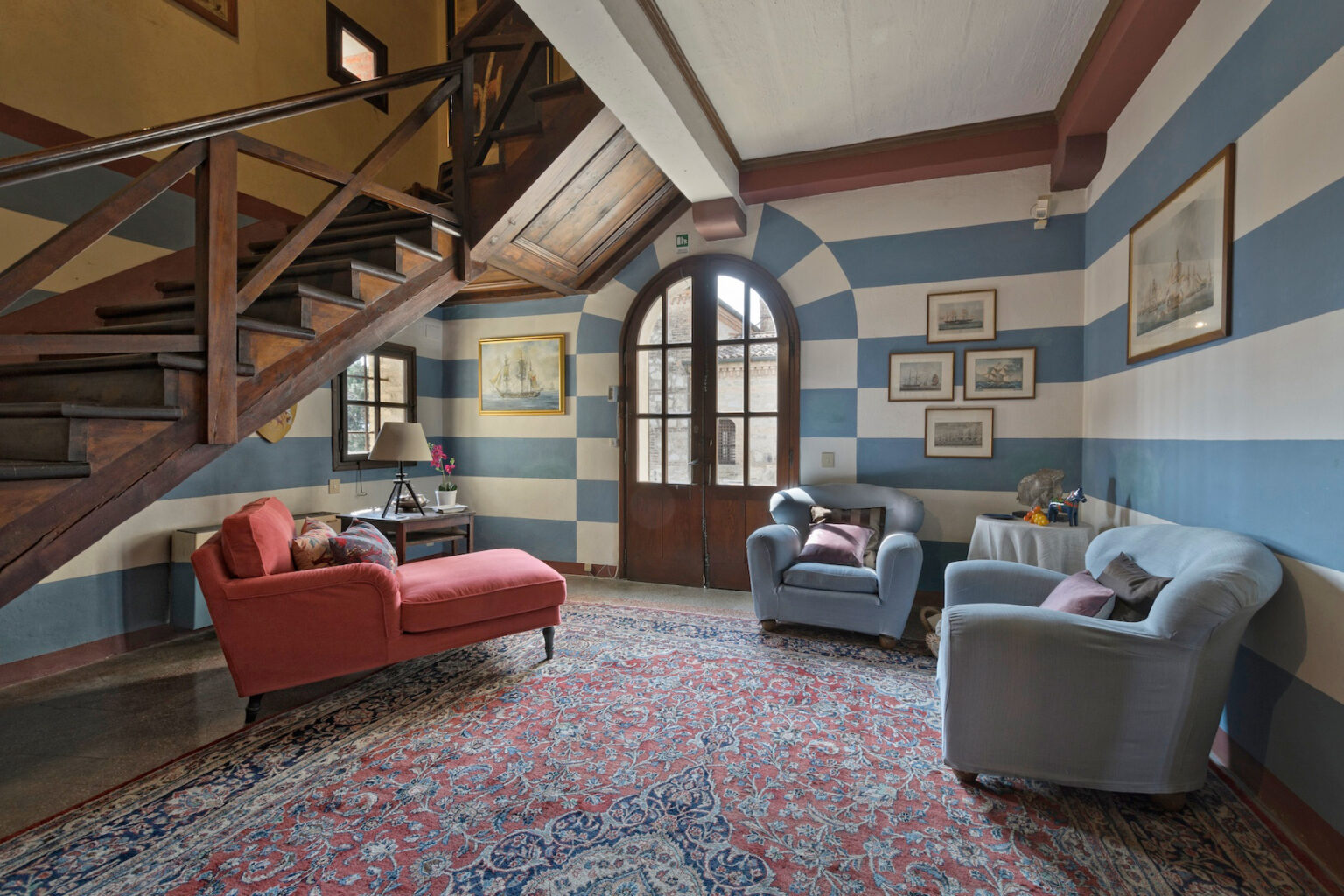 Cozy living room featuring striped blue and beige walls, a wooden staircase, vintage furniture including a red sofa and a blue armchair, and decorative artwork, illuminated by natural light from large windows. | Only on SopranoVillas