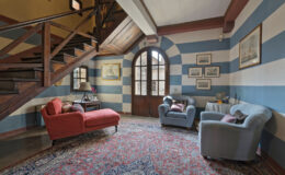 Cozy living room featuring striped blue and beige walls, a wooden staircase, vintage furniture including a red sofa and a blue armchair, and decorative artwork, illuminated by natural light from large windows.