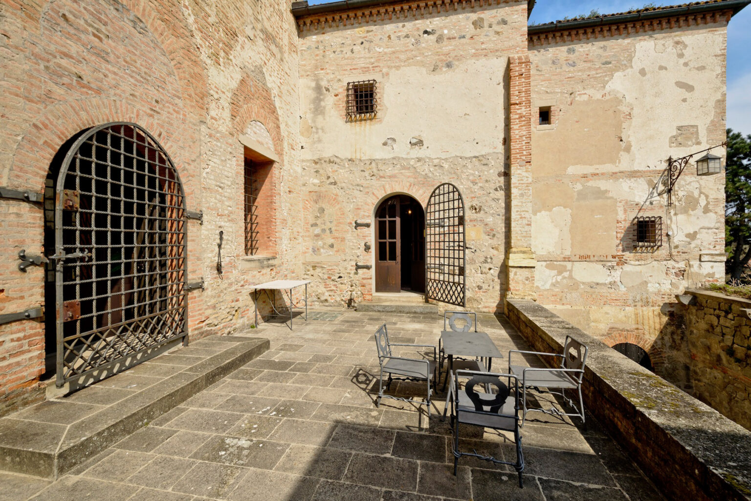 Historic castle courtyard featuring stone walls, arched doorways, and wrought iron gates, with a small table and chairs for outdoor seating. Ideal for showcasing medieval architecture and outdoor dining spaces. | Only on SopranoVillas