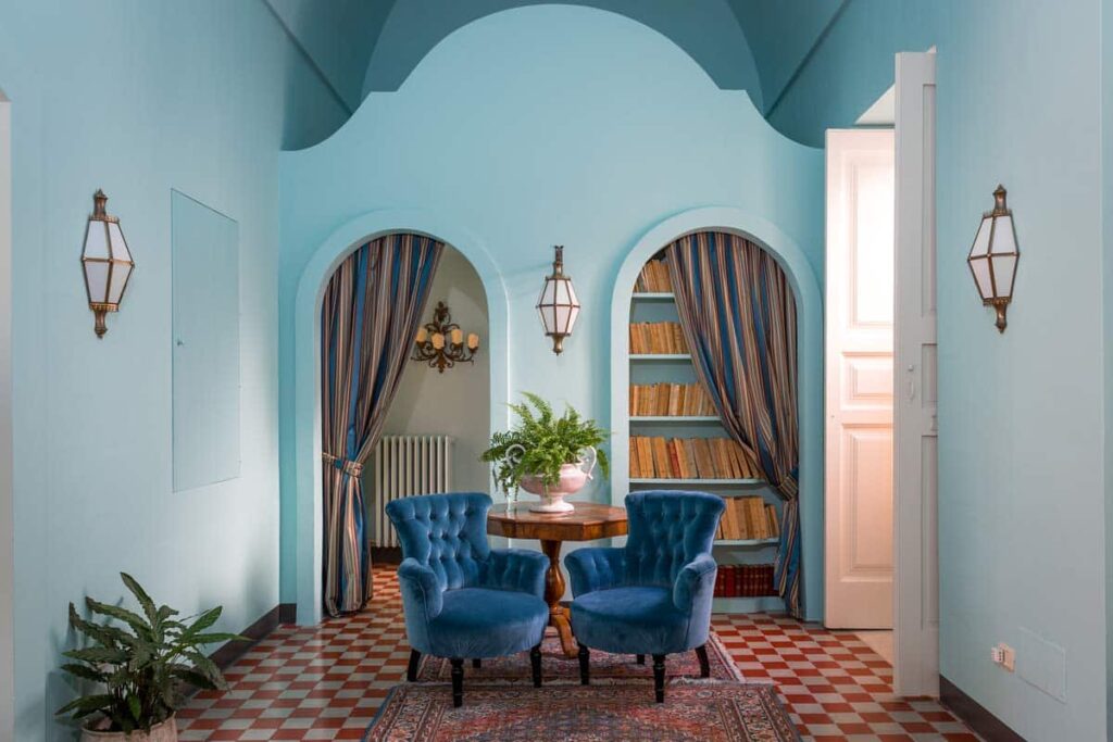 Interior view of a stylish room featuring two blue velvet chairs, a round table with a potted plant, and arched doorways with elegant curtains. The walls are painted a soothing light blue, complemented by decorative lanterns and patterned tile flooring. | Only on SopranoVillas