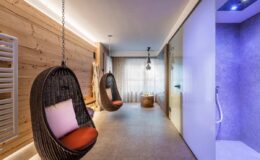 Modern relaxation space featuring two hanging wicker chairs with orange cushions, warm wooden accents, ambient lighting, and a sleek shower area in a contemporary setting.