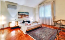 Cozy bedroom featuring a wooden floor, large window with sheer curtains, and a decorative area rug. The room includes a minimalist bed with white linens, a bedside table with lamps, and artistic wall decor for a tranquil atmosphere.