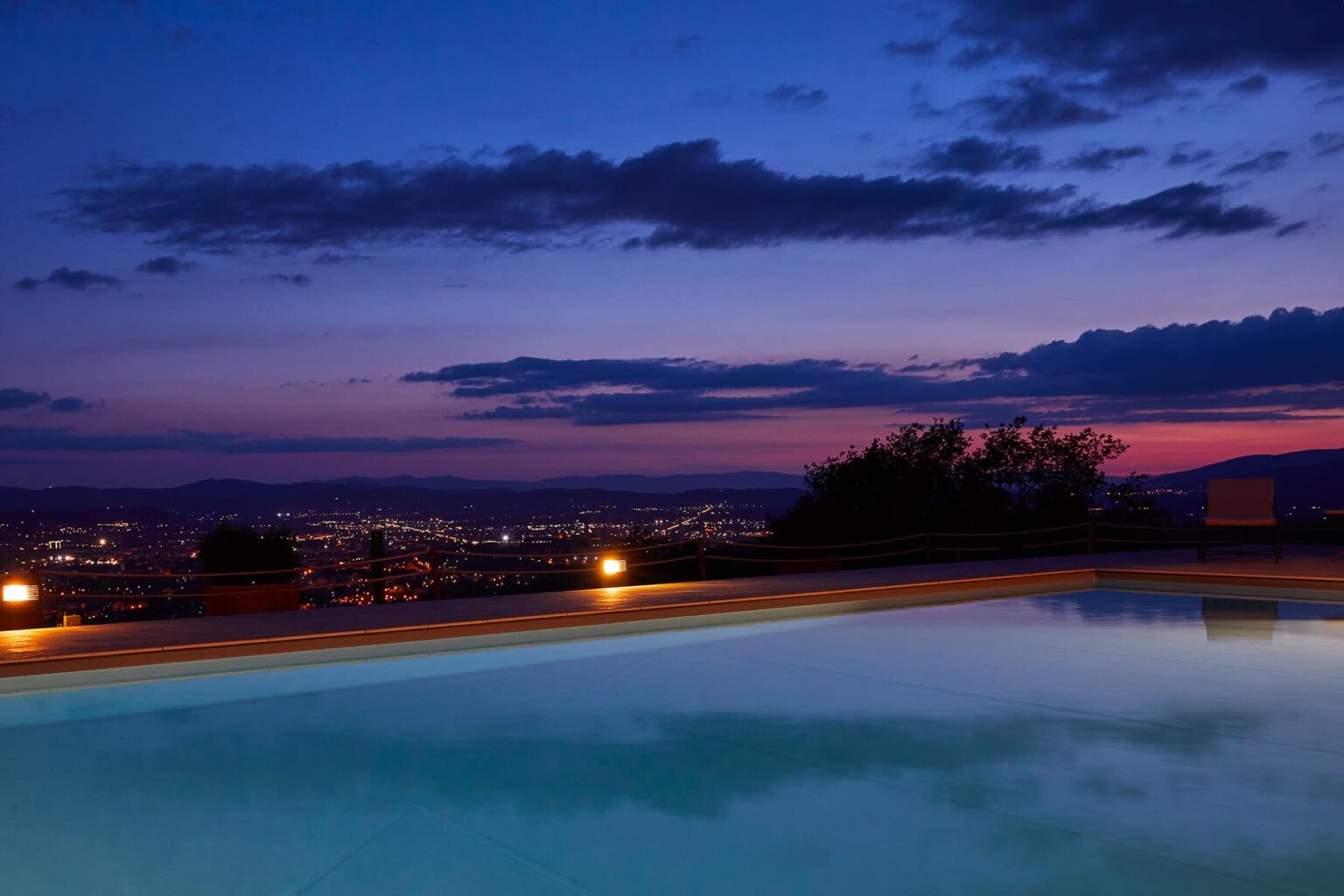 Scenic view of a tranquil pool at dusk, surrounded by city lights and a colorful sunset sky, highlighting relaxation and luxury. | Only on SopranoVillas