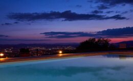 Scenic view of a tranquil pool at dusk, surrounded by city lights and a colorful sunset sky, highlighting relaxation and luxury.