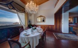 Elegant dining room with a round table set for a meal, featuring a chandelier and large window offering scenic views. The decor includes classic wooden elements and tasteful furnishings, creating a sophisticated atmosphere.
