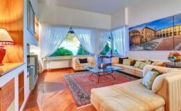 Bright and spacious living room featuring a modern sofa, coffee table, and large windows with sheer curtains, complemented by a decorative rug and a wall art piece depicting an architectural scene.