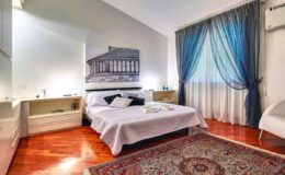 Modern bedroom with a large bed, stylish decor, and natural light from a window draped with blue curtains. The room features wooden flooring, a decorative rug, and minimalist furnishings, creating a cozy and inviting atmosphere.
