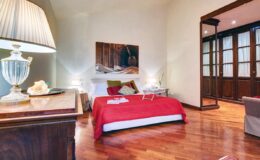 Cozy bedroom featuring a red bedspread, wooden flooring, and a stylish lamp. The room has elegant wooden cabinetry and a large window, creating a warm and inviting atmosphere. Ideal for relaxation and comfort.