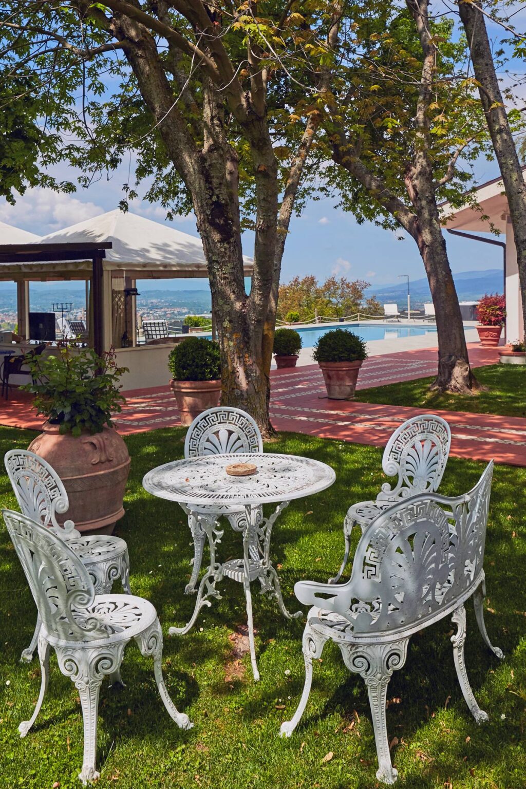Outdoor patio area featuring a white wrought iron table and chairs, surrounded by lush greenery and potted plants, with a swimming pool and scenic views in the background. Perfect for relaxation and entertaining guests. | Only on SopranoVillas