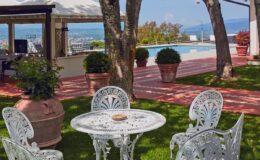 Outdoor patio area featuring a white wrought iron table and chairs, surrounded by lush greenery and potted plants, with a swimming pool and scenic views in the background. Perfect for relaxation and entertaining guests.