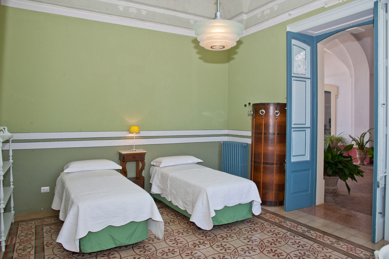Cozy bedroom featuring two twin beds with white linens and green accents, set against a soothing green wall. The room includes a bedside table, a wooden wardrobe, and a door leading to a balcony, complemented by natural light and indoor plants. | Only on SopranoVillas