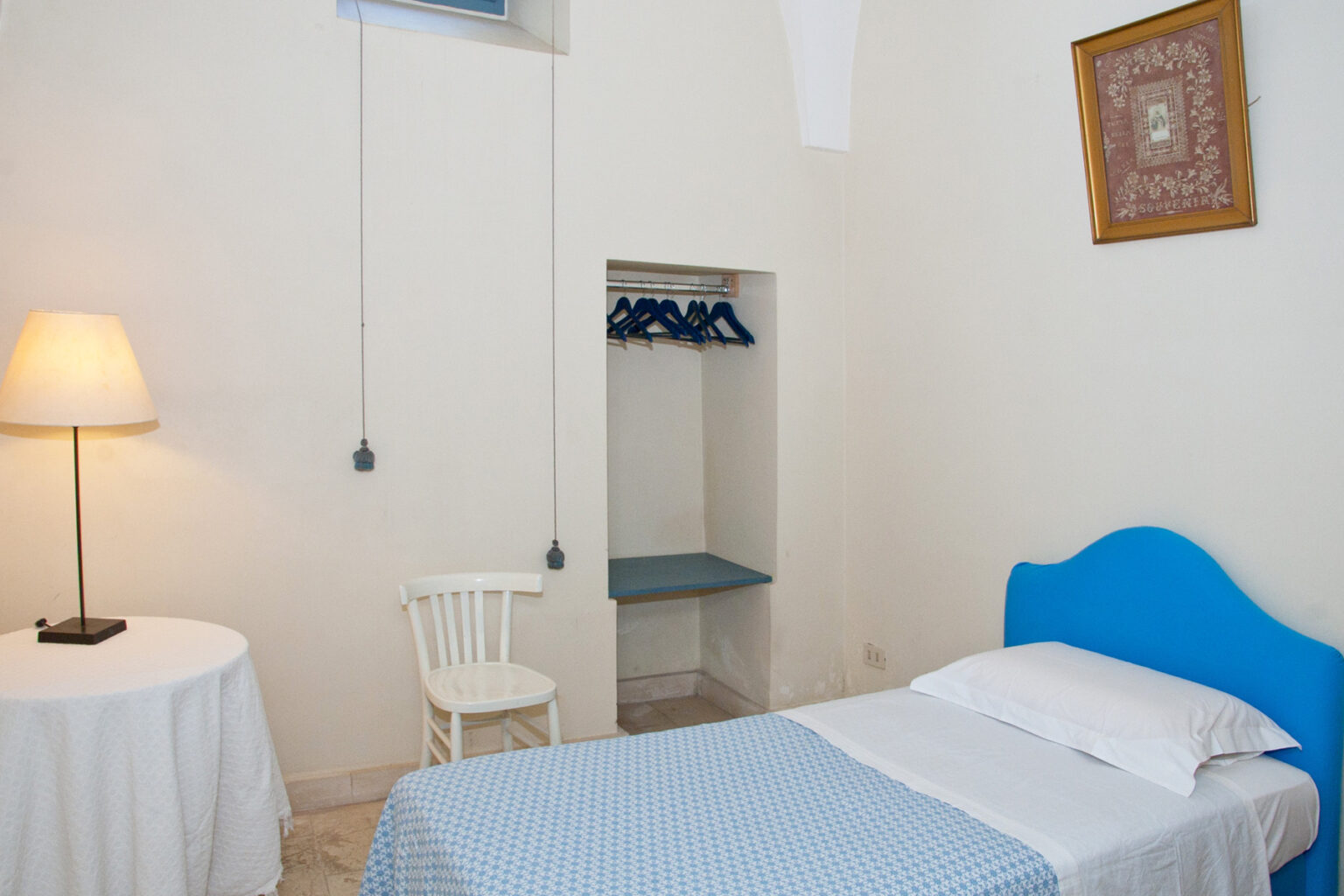 Cozy bedroom featuring a single bed with blue headboard, a white table and chair, and a closet area with hanging space. Soft lighting creates a warm atmosphere, ideal for relaxation. | Only on SopranoVillas