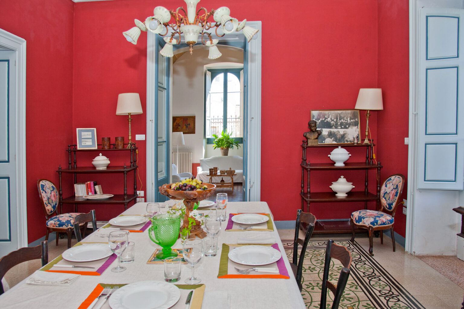 Brightly decorated dining room with a red accent wall, a large table set for a meal, colorful tableware, and decorative furnishings. Features include a chandelier, blue doors, and a view of a sunny room with green plants. | Only on SopranoVillas