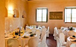 Elegant dining room set for an event, featuring round tables with white tablecloths, wine glasses, and decorative centerpieces, complemented by warm-toned walls and natural light from large windows.