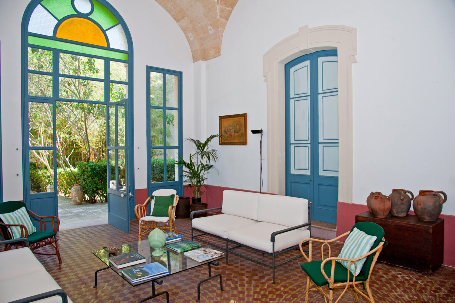 Bright and airy living room featuring large blue-framed windows, a light-colored sofa, and wooden chairs. Decorative elements include potted plants and clay pots, complemented by a patterned tile floor. | Only on SopranoVillas