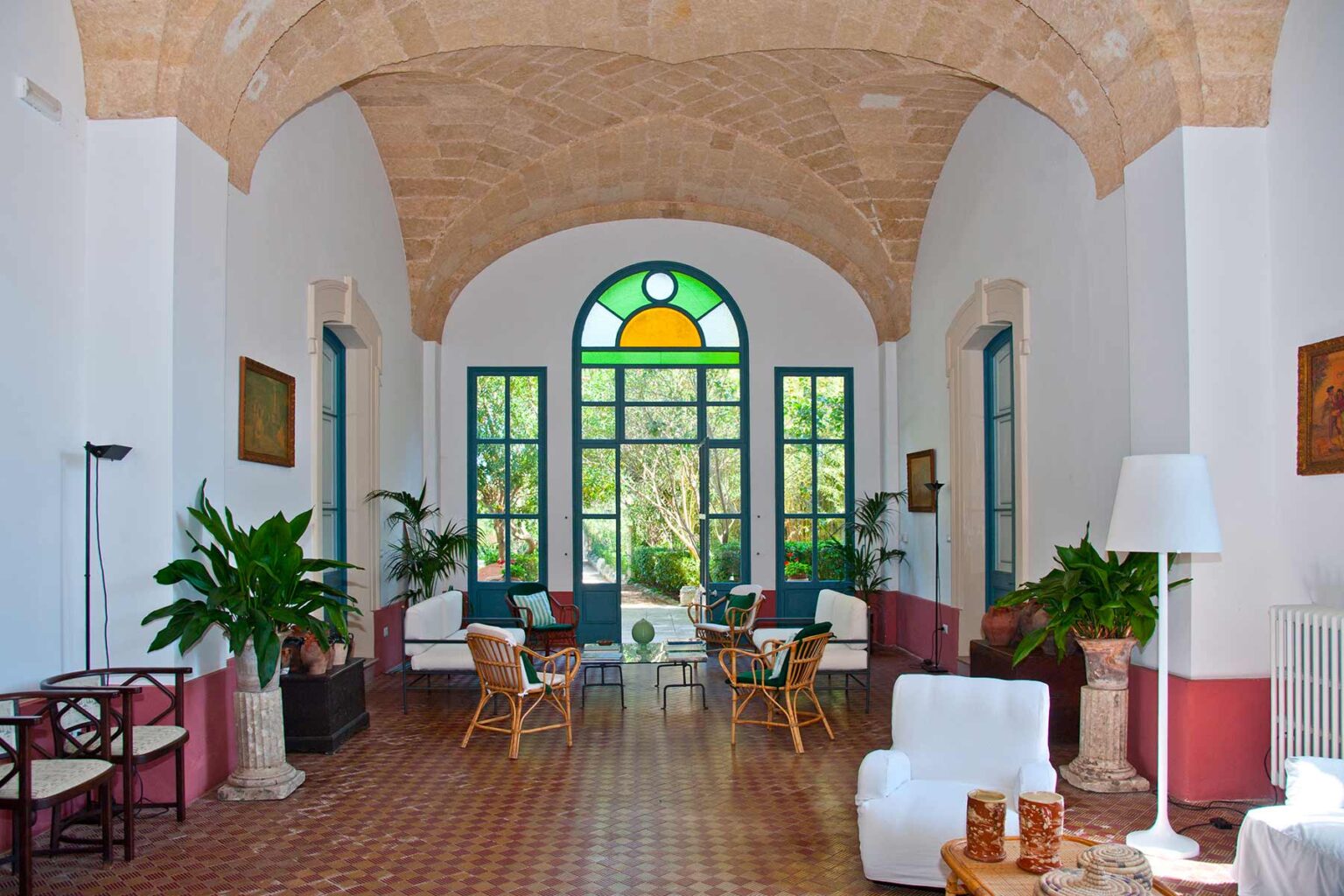 Spacious interior of a stylish room featuring arched ceilings, large windows with colorful stained glass, and comfortable seating arrangements surrounded by greenery. | Only on SopranoVillas
