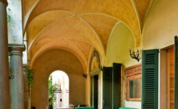 Elegant arched corridor with warm yellow walls, featuring green seating and open shutters, surrounded by lush greenery, creating a serene and inviting atmosphere.