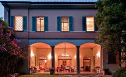 Elegant two-story house with blue shutters and arched columns, featuring a cozy dining area visible through large windows, surrounded by lush greenery and flowering plants at dusk.