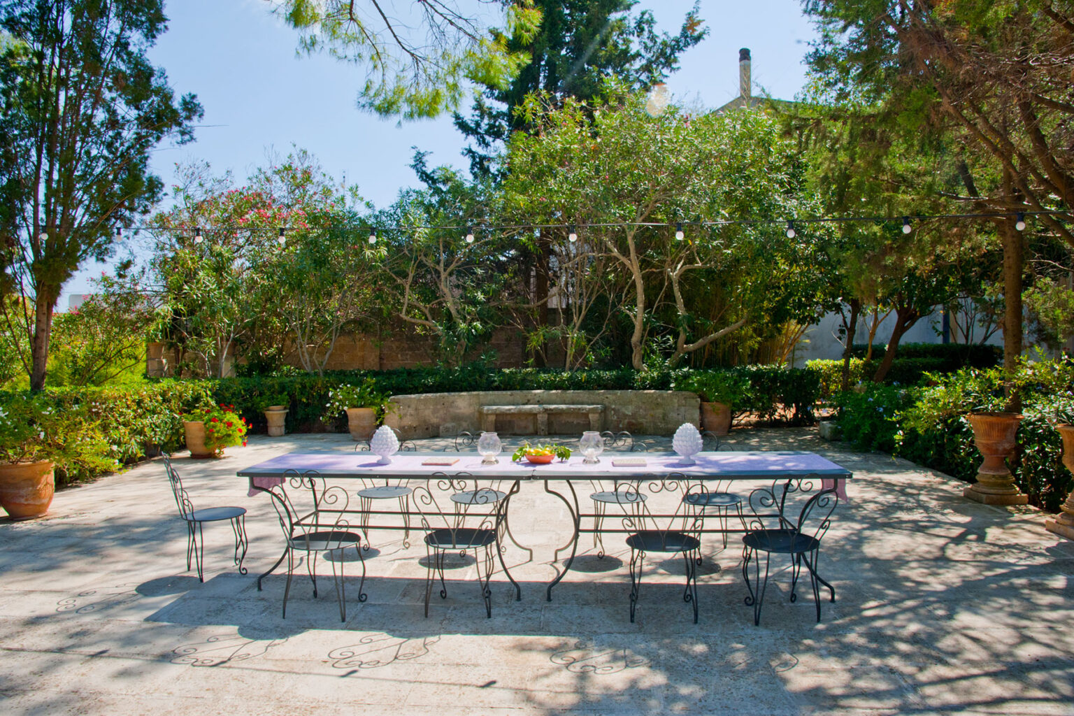 Outdoor dining area featuring a long table set for a meal, surrounded by lush greenery and potted plants. Bright sunlight filters through the trees, creating a serene ambiance perfect for gatherings. | Only on SopranoVillas