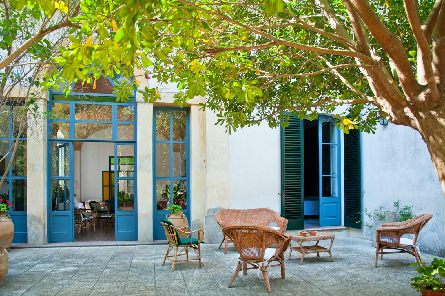 Charming outdoor seating area with wicker furniture under a lush tree, adjacent to a bright, airy room featuring large blue windows and a welcoming interior. Ideal for relaxing or entertaining in a tranquil garden setting. | Only on SopranoVillas
