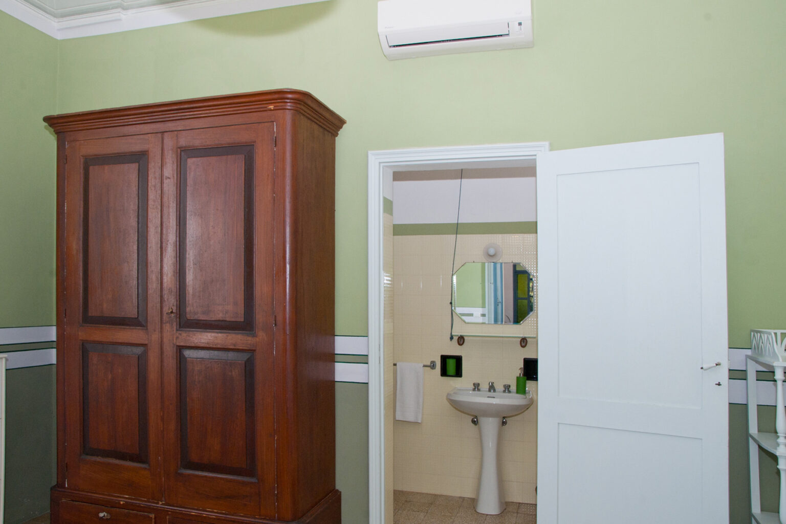 Interior view of a room featuring a wooden wardrobe, a white door leading to a bathroom with a sink and mirror, and an air conditioning unit on the wall, all set against a light green background. | Only on SopranoVillas