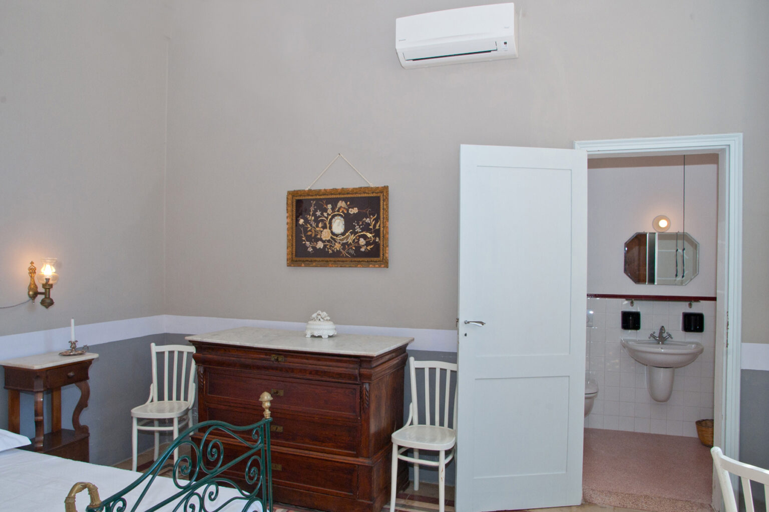 Cozy bedroom interior featuring a wooden dresser, two white chairs, and a decorative painting on the wall. A bathroom doorway is visible, showcasing a sink and mirror. The room is well-lit with an air conditioning unit mounted on the wall, enhancing comfort. | Only on SopranoVillas