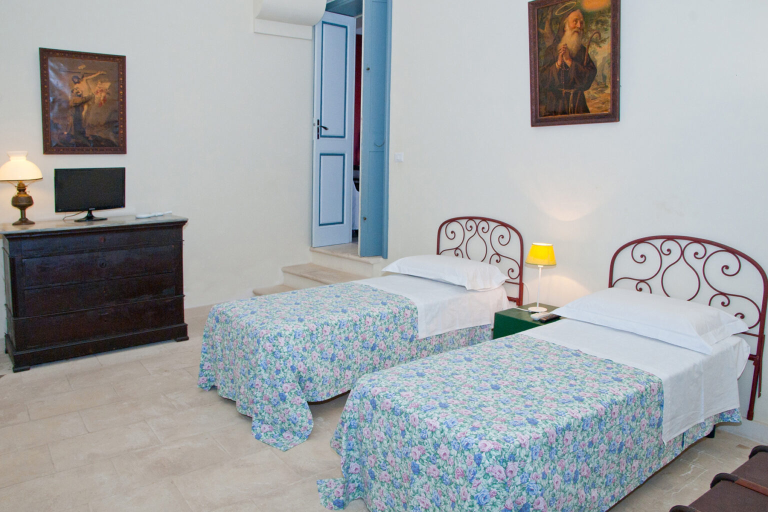 Cozy guest room featuring two twin beds with floral-patterned bedspreads, a wooden dresser, and soft lighting. The room is adorned with wall art and has a door leading to a balcony. Ideal for travelers seeking comfort and style. | Only on SopranoVillas