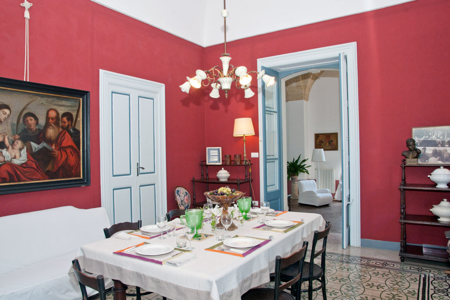 Elegant dining room featuring a white table set for a meal, surrounded by red walls and a large painting. A chandelier hangs above, and a doorway leads to a bright living space with plants. Ideal for showcasing interior design and dining decor. | Only on SopranoVillas