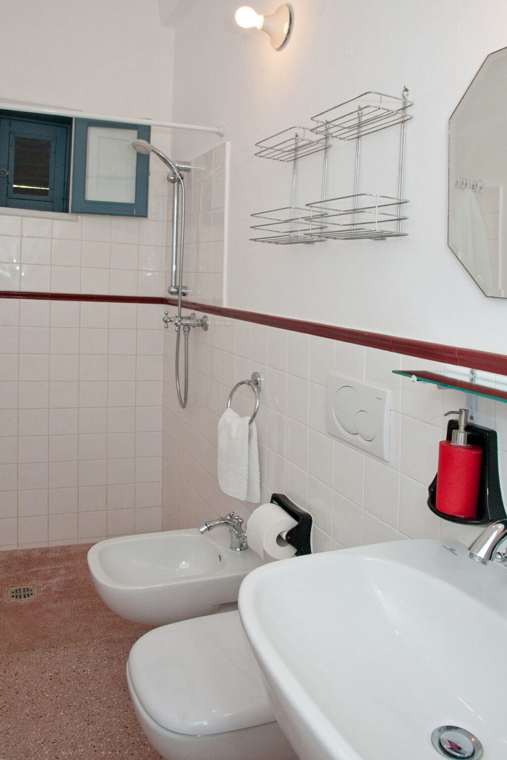 Modern bathroom featuring a shower with a handheld fixture, white tiled walls, and a towel rack. The design includes a mirror, a glass shelf with toiletries, and a red accent piece for added style. | Only on SopranoVillas