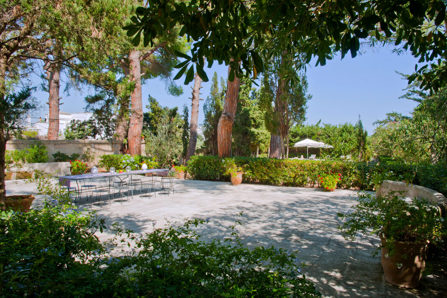 Lush garden landscape featuring tall trees, green foliage, and a stone patio set with outdoor furniture, ideal for relaxation and gatherings. | Only on SopranoVillas