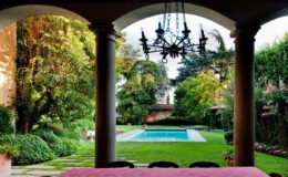View of a landscaped garden with a swimming pool, framed by elegant columns and a chandelier overhead, showcasing lush greenery and vibrant flowers.