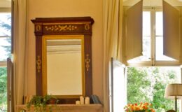 Elegant interior featuring a vintage wooden mirror above a mantel, with natural light streaming through open windows. The decor includes candles and a vase of fresh flowers, enhancing the room's warm ambiance.