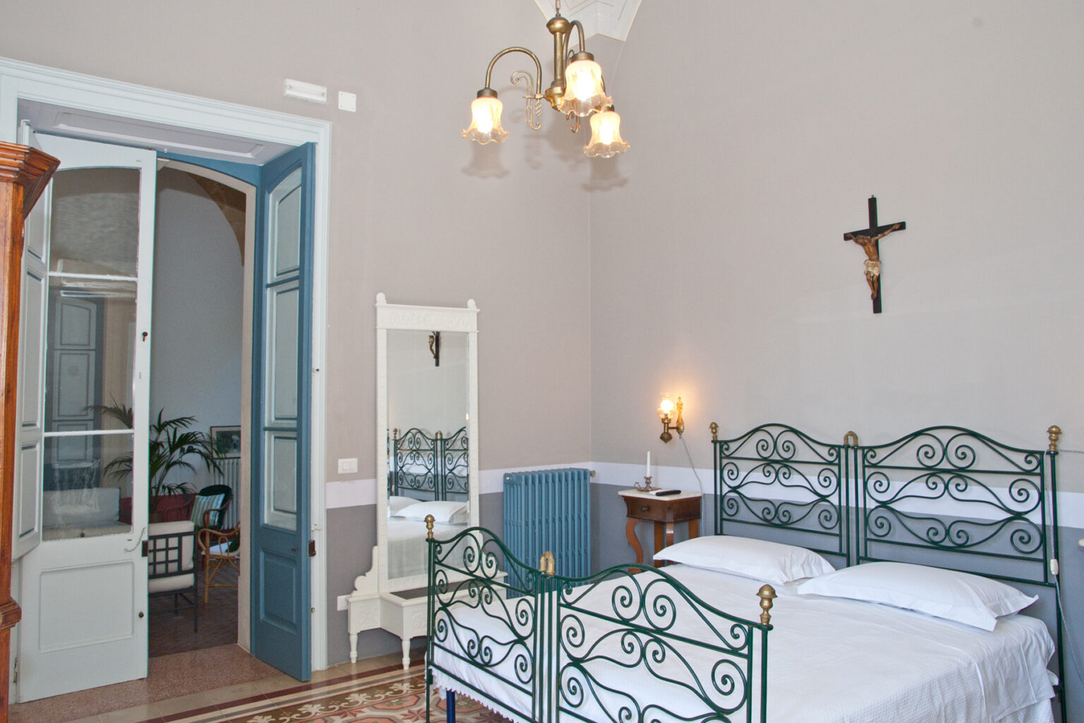 Cozy bedroom featuring vintage iron beds, a wooden dresser, and a mirror, with a light-colored wall and a doorway leading to a bright sitting area. The room is illuminated by a stylish chandelier and has a decorative cross on the wall. | Only on SopranoVillas