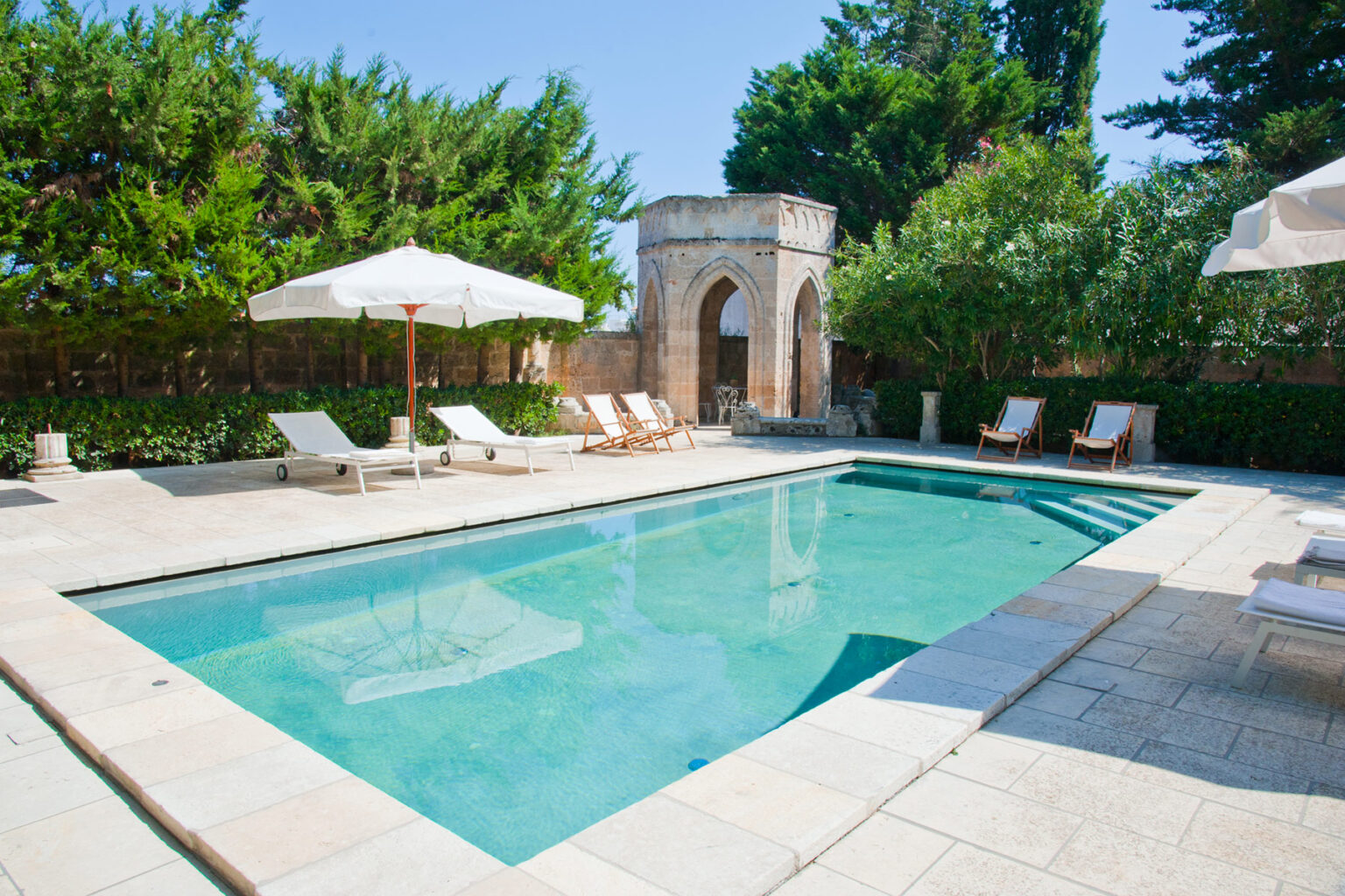 Luxury outdoor swimming pool surrounded by lounge chairs and sun umbrellas in a serene garden setting. | Only on SopranoVillas