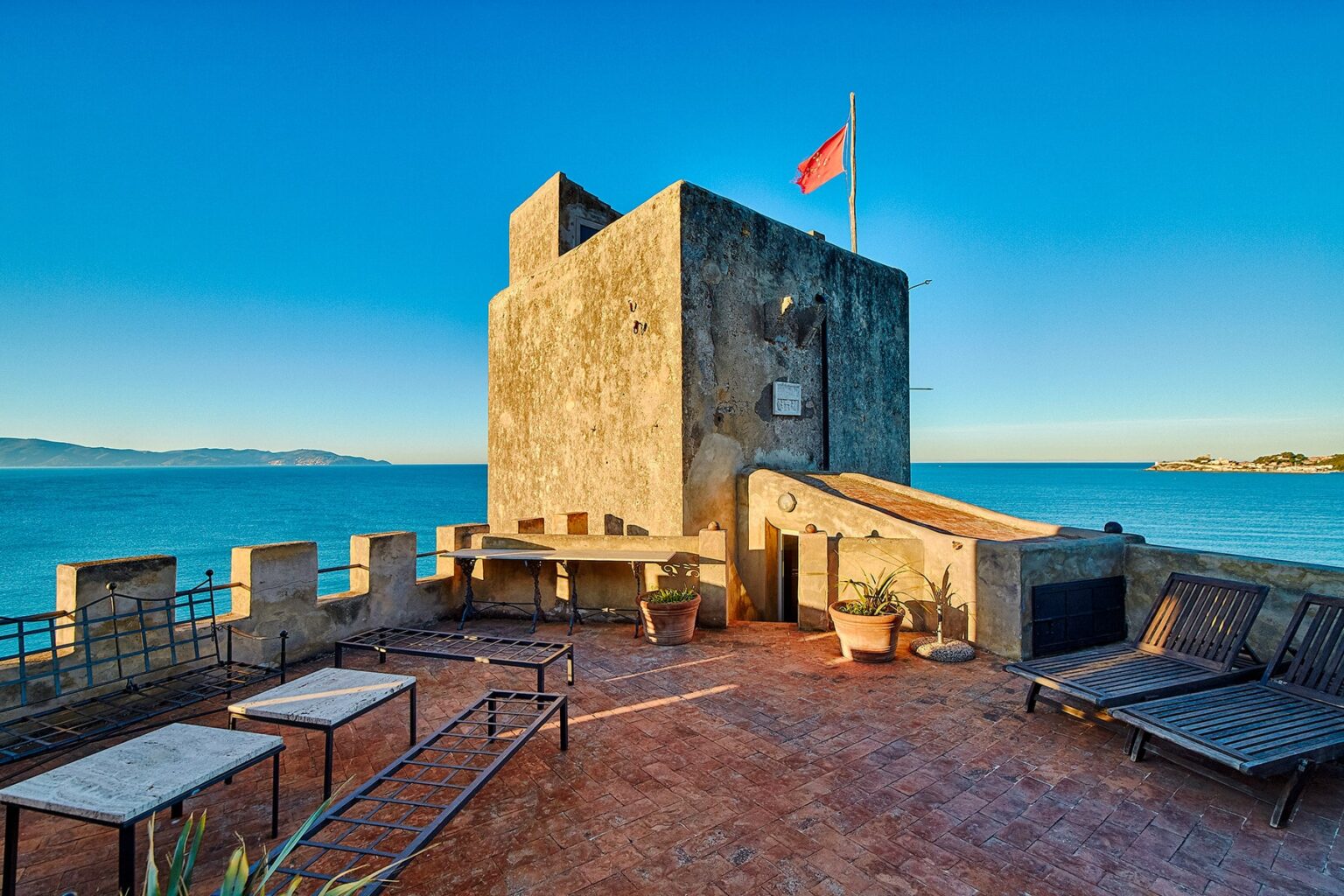 Historic coastal tower with a flag, overlooking the sea, featuring a terrace with lounge chairs in the foreground and a clear blue sky. | Only on SopranoVillas