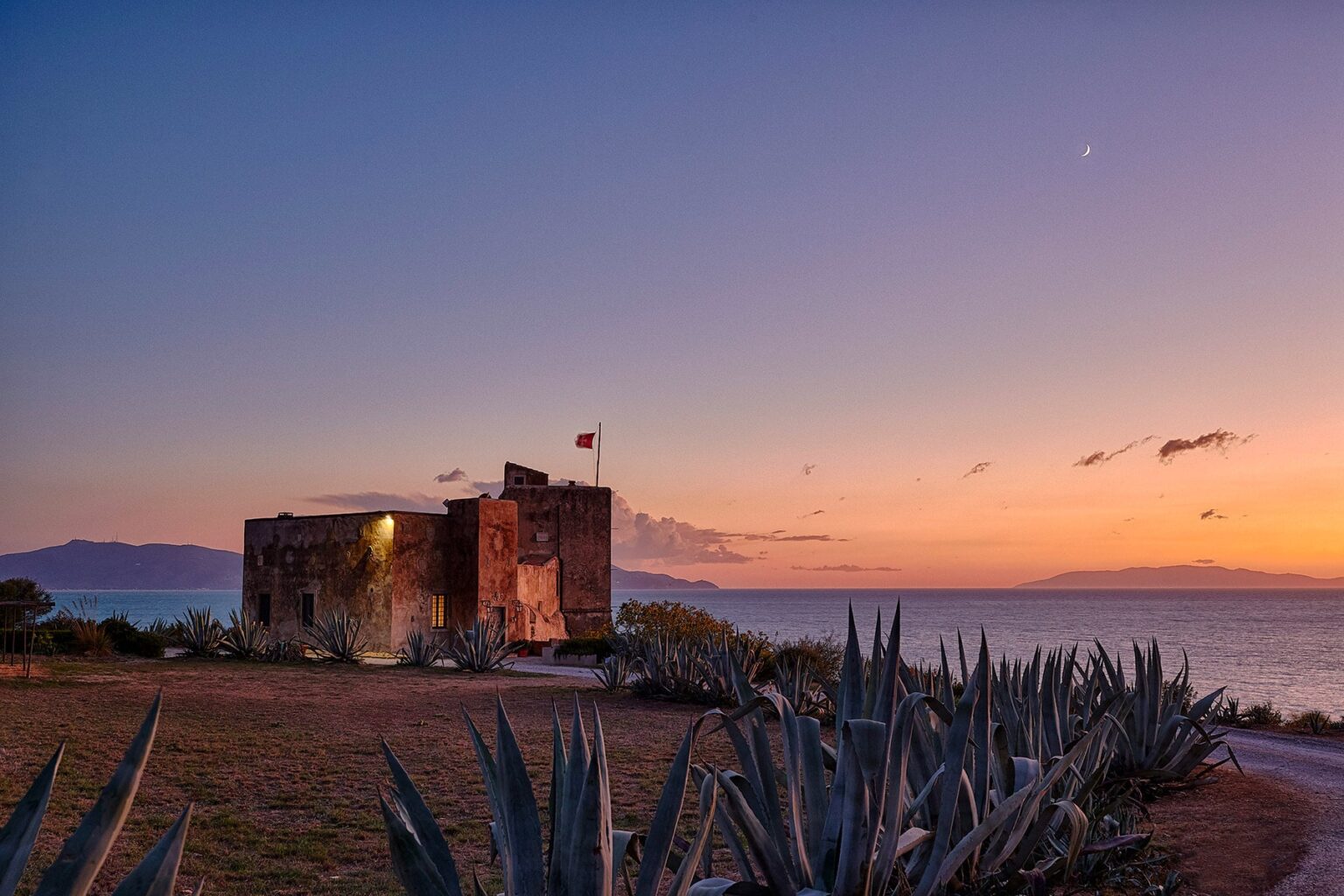 Historic stone building by the sea at sunset, featuring a flag and surrounded by agave plants, with a colorful sky in the background. | Only on SopranoVillas