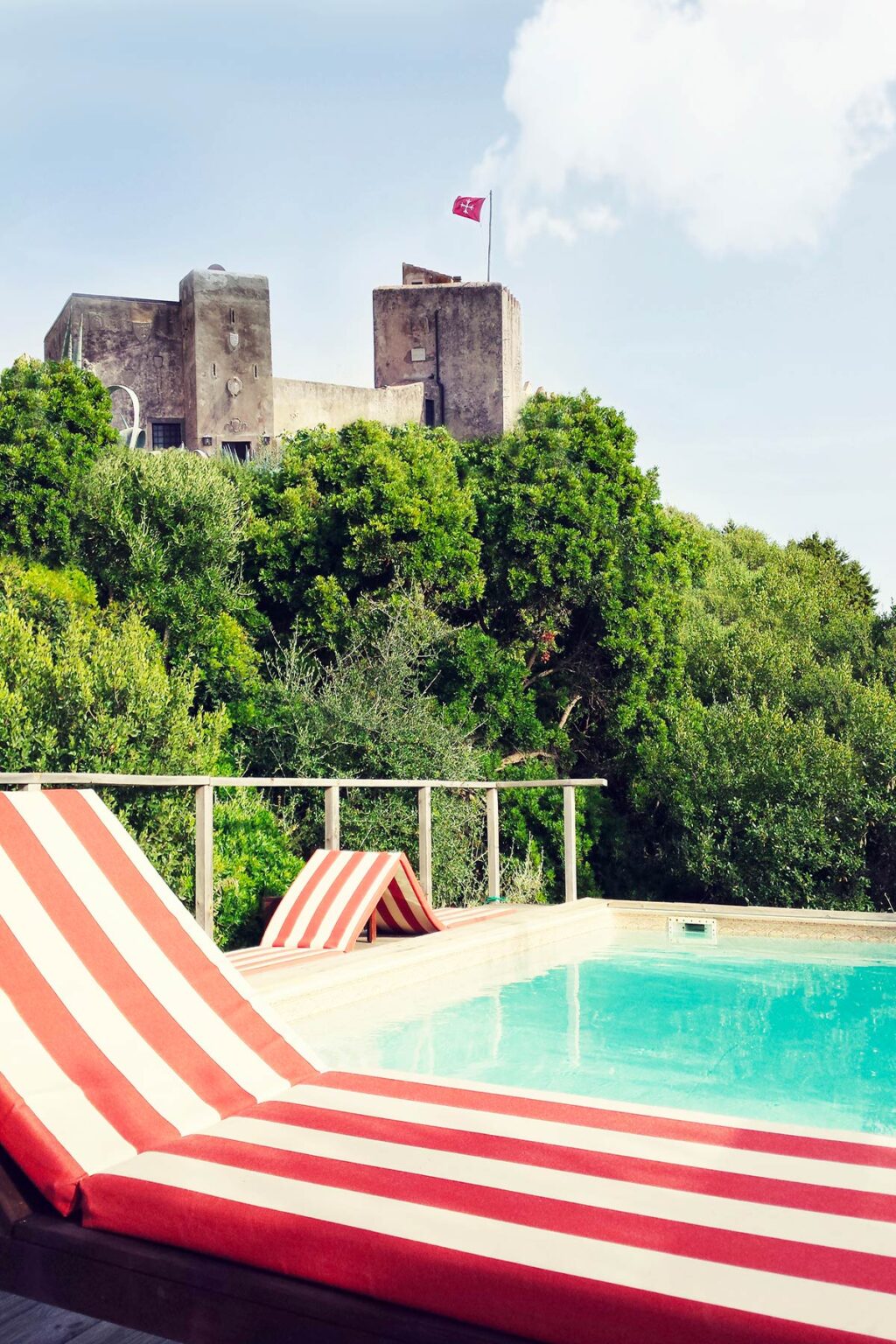 Poolside view with striped lounge chairs overlooking lush greenery and a distant historic building, ideal for relaxation and scenic enjoyment. | Only on SopranoVillas