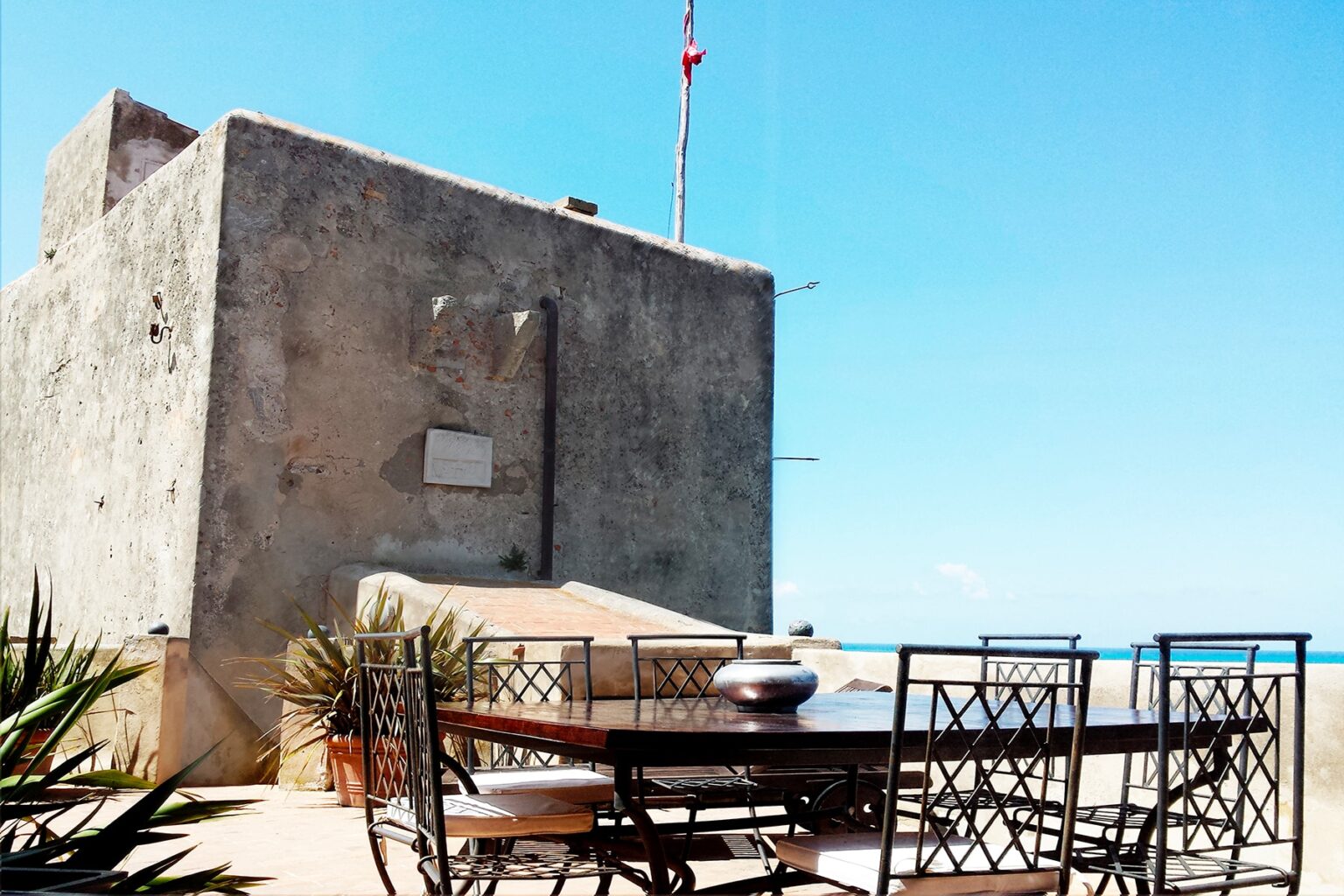 Outdoor dining area with a wooden table and black metal chairs, set against a rustic stone building and clear blue sky, ideal for enjoying meals in a scenic location. | Only on SopranoVillas