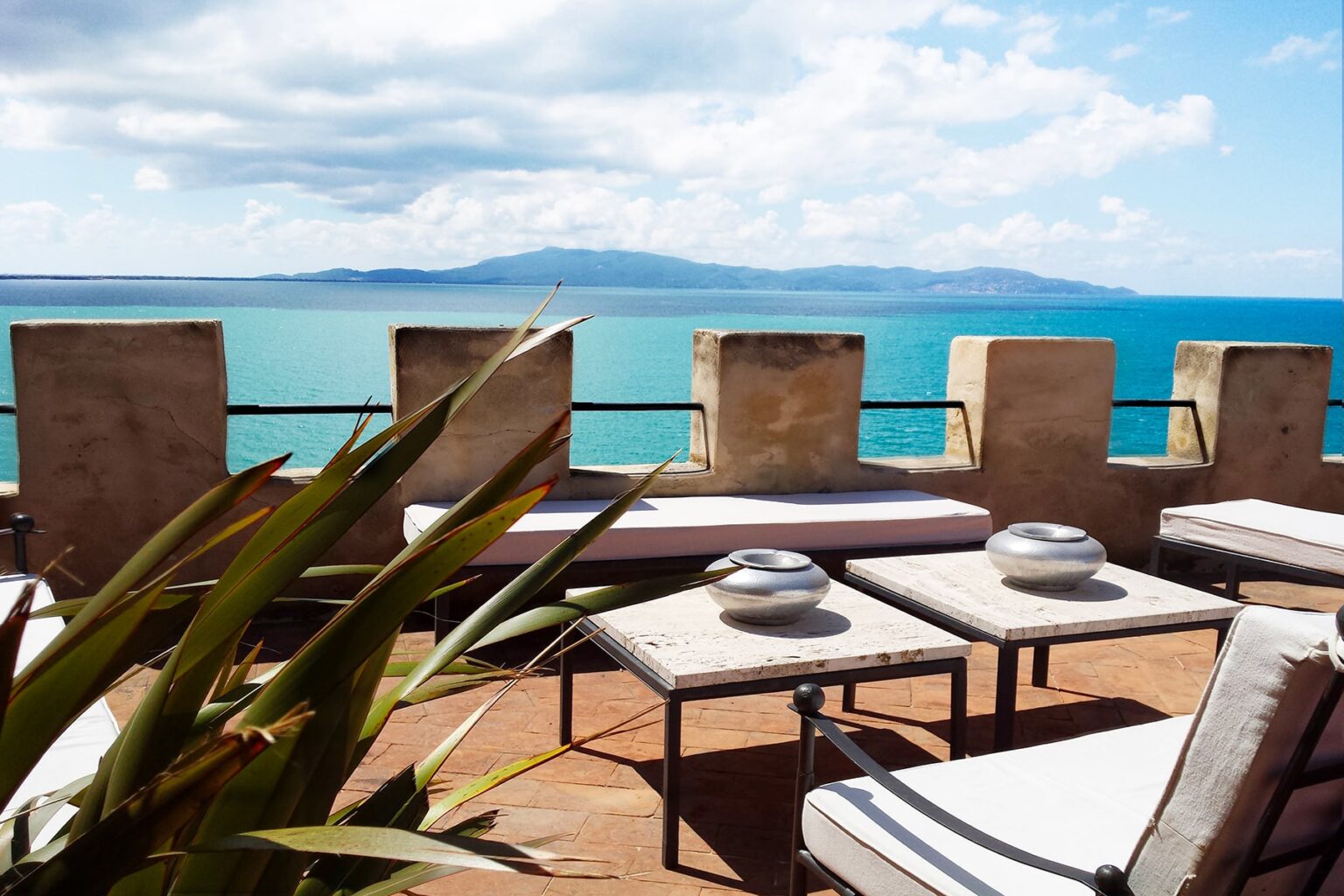 Scenic ocean view from a terrace featuring modern outdoor furniture, lush greenery, and a clear blue sky with clouds. Ideal for relaxation and enjoying coastal landscapes. | Only on SopranoVillas