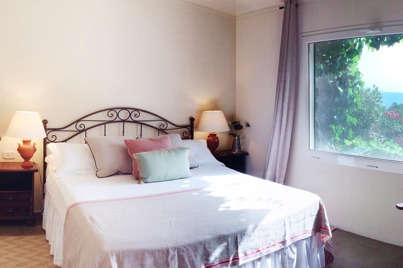 Cozy bedroom featuring a wrought iron bed with white linens, decorative pillows, bedside lamps, and a large window with a view of greenery. Ideal for relaxation and comfort. | Only on SopranoVillas