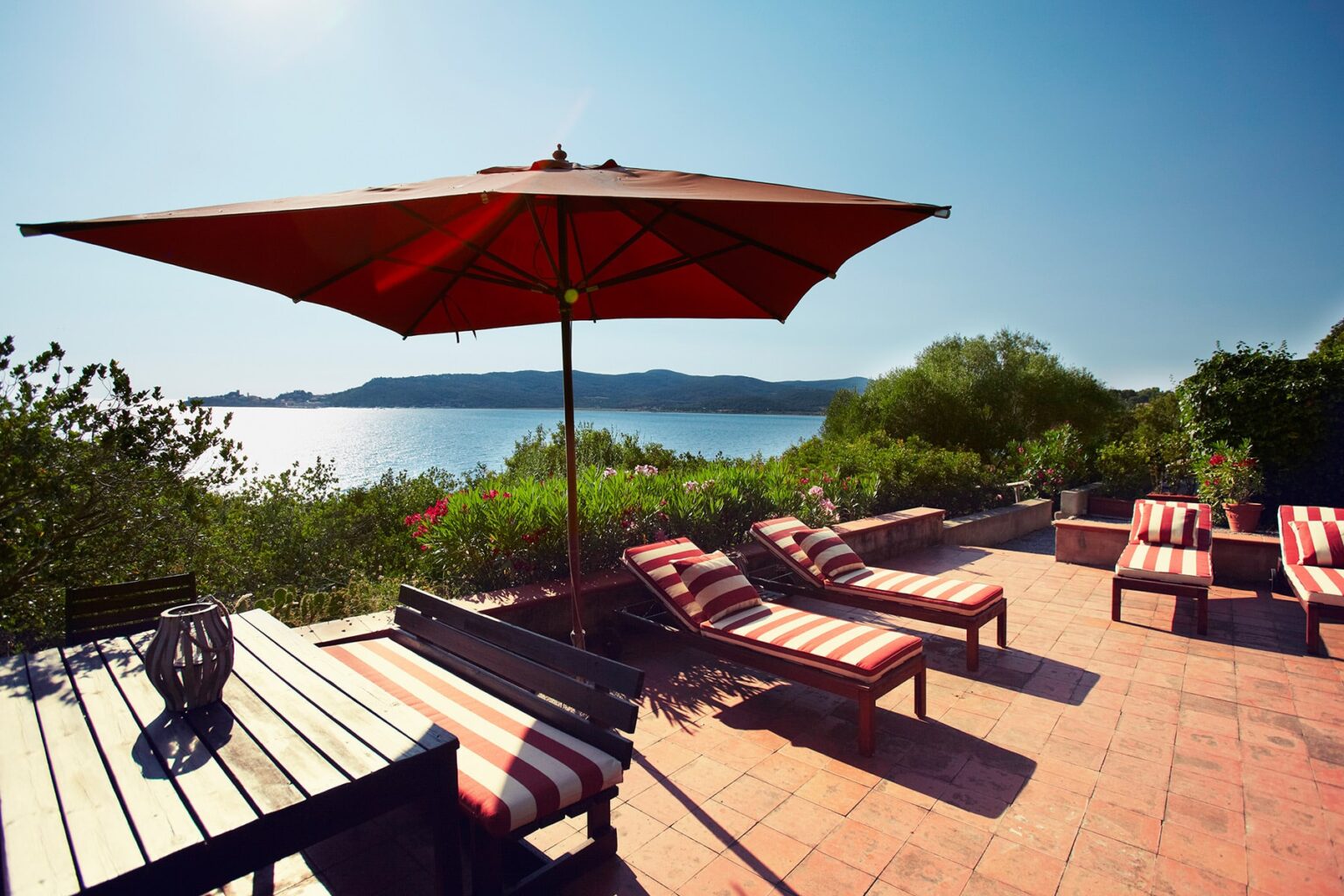 Outdoor patio with striped lounge chairs and a large red umbrella, overlooking a serene lake and lush greenery under a clear blue sky. | Only on SopranoVillas