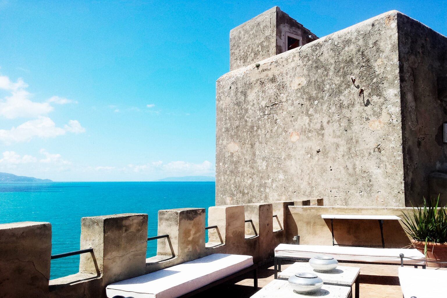 Scenic view of a coastal terrace featuring a stone wall and seating area, overlooking a vibrant blue sea under a clear sky. | Only on SopranoVillas