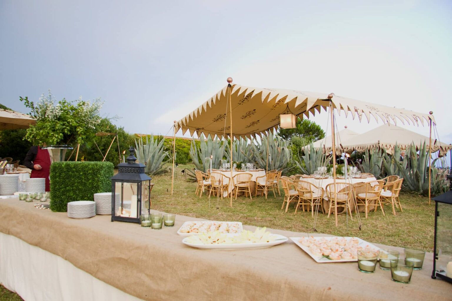Outdoor event setup featuring elegant wooden dining chairs and tables beneath a decorative tent, surrounded by lush greenery and decorative lanterns. Ideal for weddings, parties, or gatherings in a natural setting. | Only on SopranoVillas